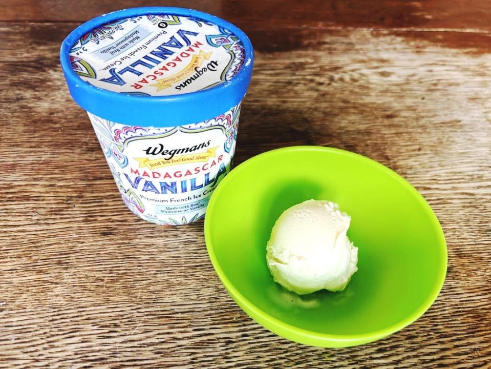 A white pint of ice cream with red, green, and blue detailing and Wegmans logo on the front. The pint sits next to a green bowl holding a scoop of vanilla ice cream