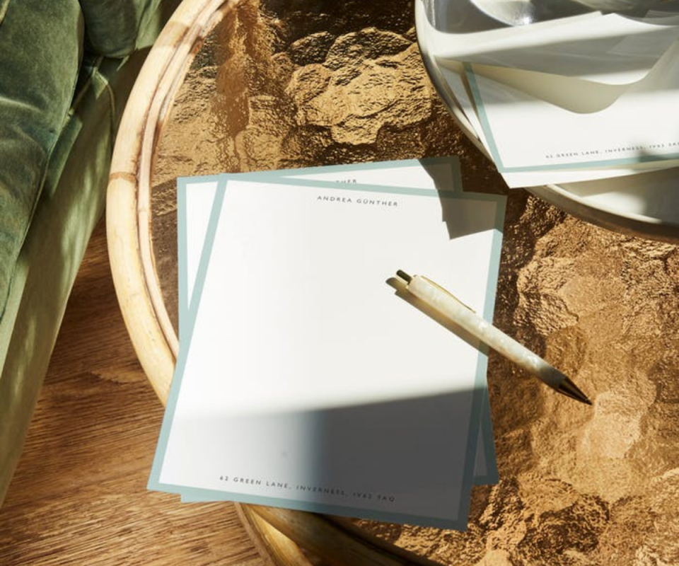 White and mint-trimmed writing paper with a pearl pen ontop on an orange-stained glass table.