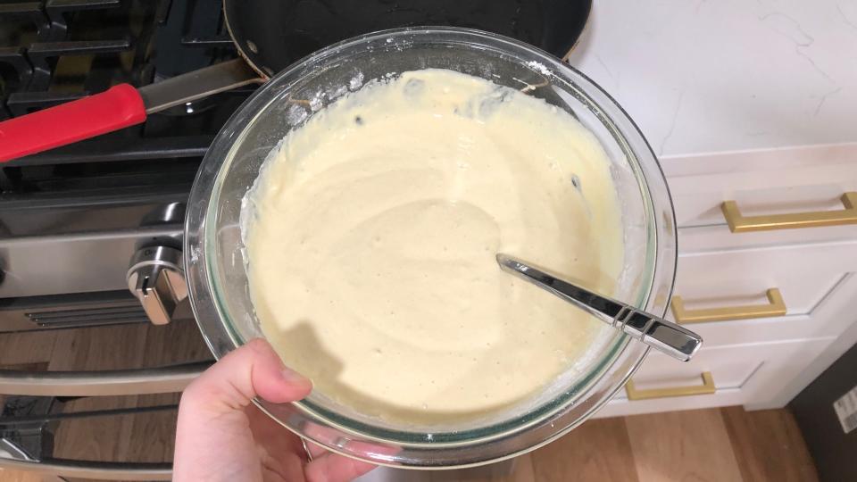 A hand holding a clear bowl filled with pancake batter. There is also a silver spoon in the bowl.