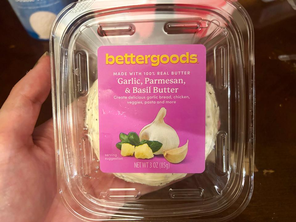 A hand holds a container of butter with a pink label with "Bettergoods garlic, Parmesan, and basil butter" text on it
