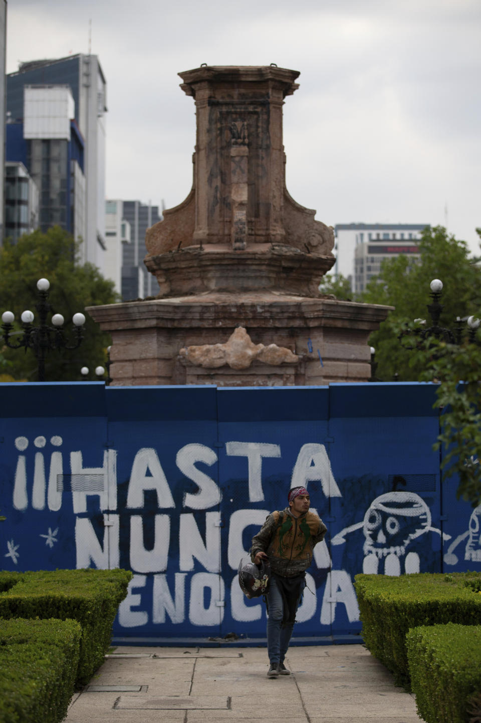 A pedestrian walks past graffiti that reads in Spanish "Never again genocider" on a temporary metal barrier set up to protect the perimeter of the Christopher Columbus statue which was removed by authorities on Paseo de la Reforma in Mexico City, Monday, Oct. 12, 2020. Mexico has has a hard time dealing with the legacy of Cristopher Columbus, and officials are still being coy about why the statue was removed over the weekend. (AP Photo/Fernando Llano)