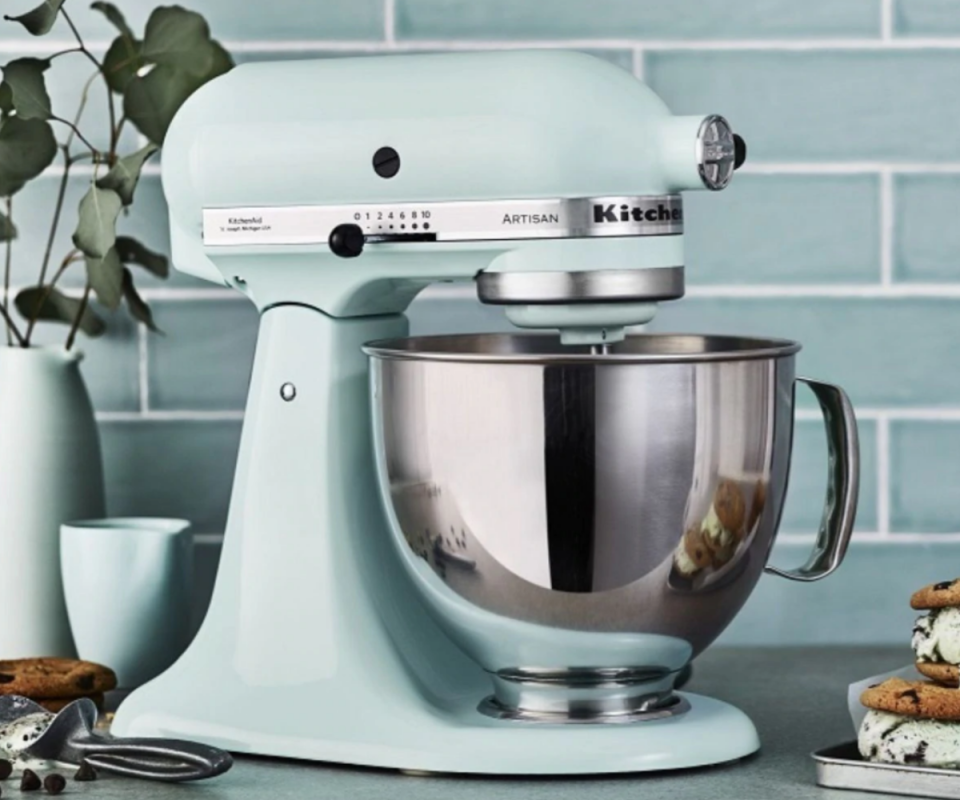 A pale blue KitchenAid mixer with a silver bowl attached sits on a grey kitchen bench with pale blue tiles behind it.