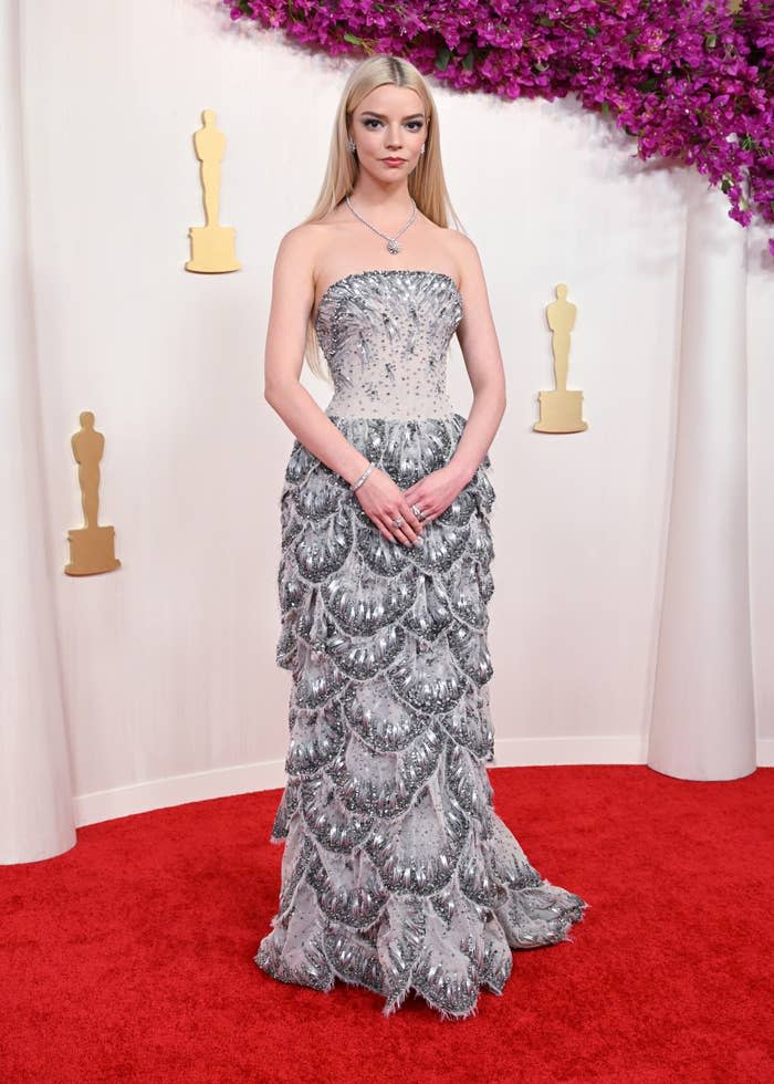 Anya Taylor-Joy poses on a red carpet in a strapless, sequined gown with a layered, scallop pattern design. Golden Oscar statues are in the background