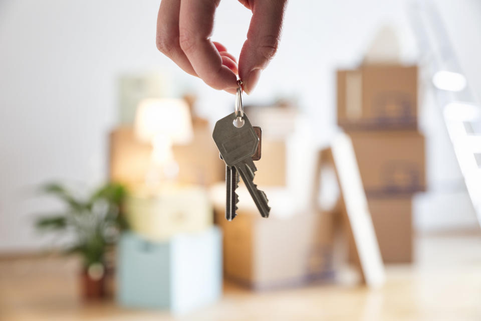 A hand holds keys in focus, with blurred moving boxes and home items in the background, symbolizing moving into a new house. No persons are visible