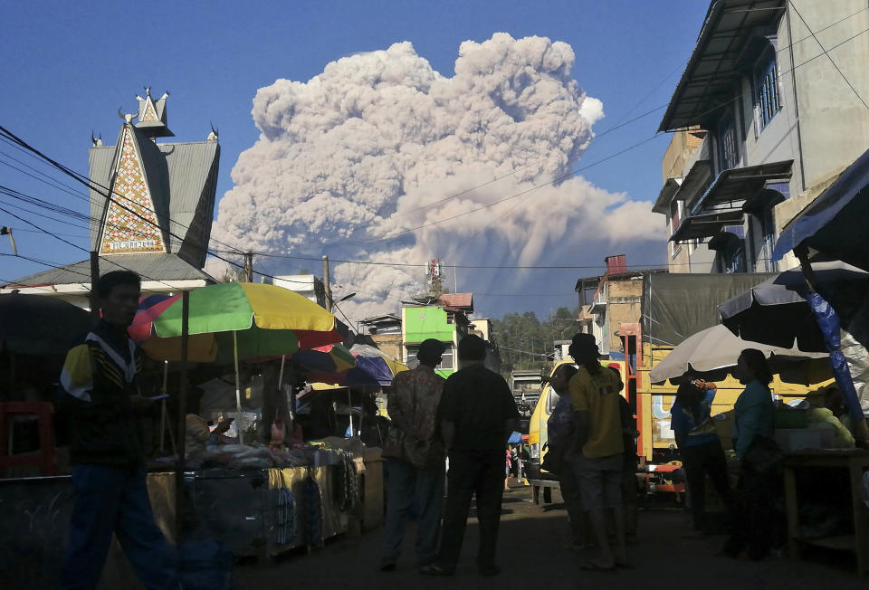 People watch as Mount Sinabung spews volcanic material during an eruption, at a market in Karo, North Sumatra, Indonesia, Tuesday, March 2, 2021. The 2,600-metre (8,530-feet) volcano erupted Tuesday, sending volcanic materials a few thousand meters into the sky and depositing ash on nearby villages. (AP Photo/Sugeng Nuryono)