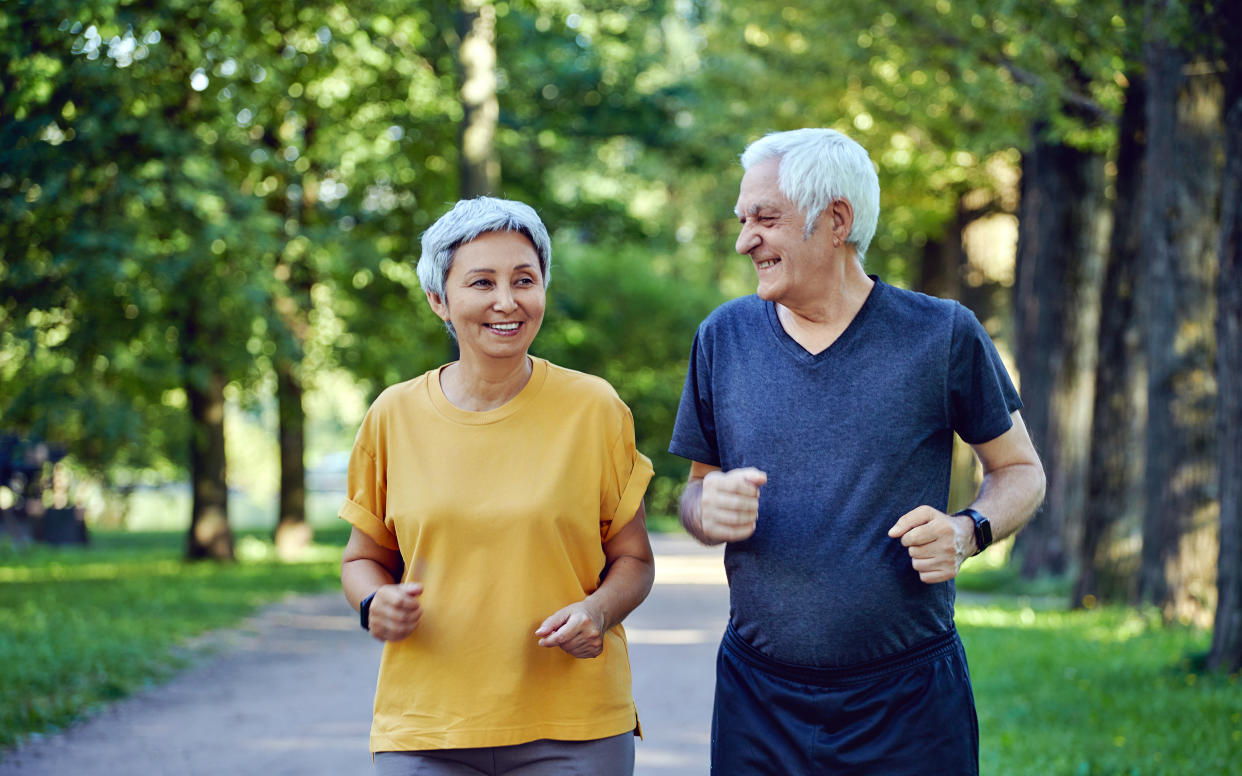 Two people with gray hair appear to jog side by side on a path through trees.