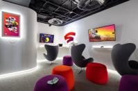 <p>Google Store Chelsea. A Stadia-themed Sandbox with wall-mounted screens, three chairs, four stools in matching purple and orange colors and a couple of controllers lying on small ledges.</p> 