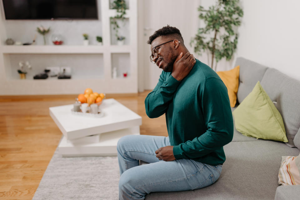This image shows a young African American man with his hand on his neck, looking uncomfortable while sitting on a sofa in a cozy living room