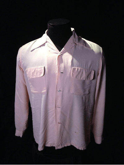 The pale pink dress shirt has wide lapels and sits on a black headless mannequin on a black background.