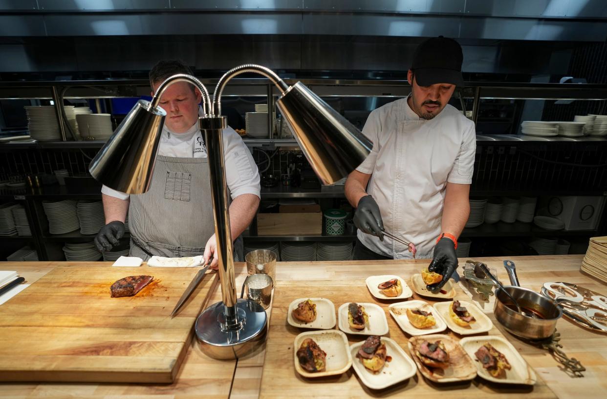 Executive chef Corey Fuller, left, and pastry chef Youssef Boudarine serve steak samples Wednesday, Jan. 17, 2024, during a preview event for Commission Row, a new restaurant, bar and event space near Gainbridge Fieldhouse in downtown Indianapolis. They served whiskey peppercorn-smoked New York strip steak on an everything-spiced popover, topped with red wine sauce and a truffle topping.