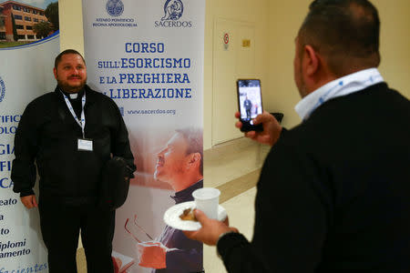 A priest poses for a picture next to a banner advertising a course for aspiring exorcists in Rome, Italy April 16, 2018. The banner reads "Course about exorcism and prayer of liberation". REUTERS/Tony Gentile