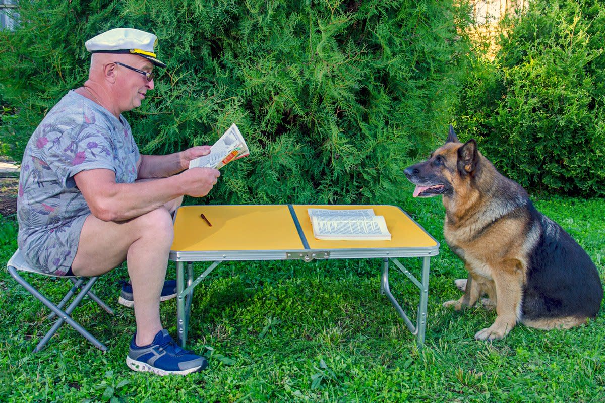 A man reads a magazine with his dog in front of him outside at a table. 