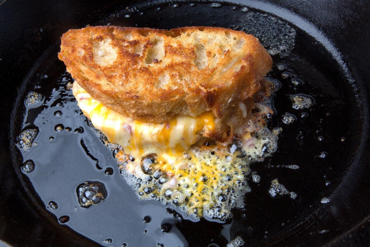 Fancy, 3 cheese grilled cheese sandwich with scallions and very thick cut baguette bread sizzling in a frying pan with melted cheese spilling out of the sandwich and bubbling in the pan.