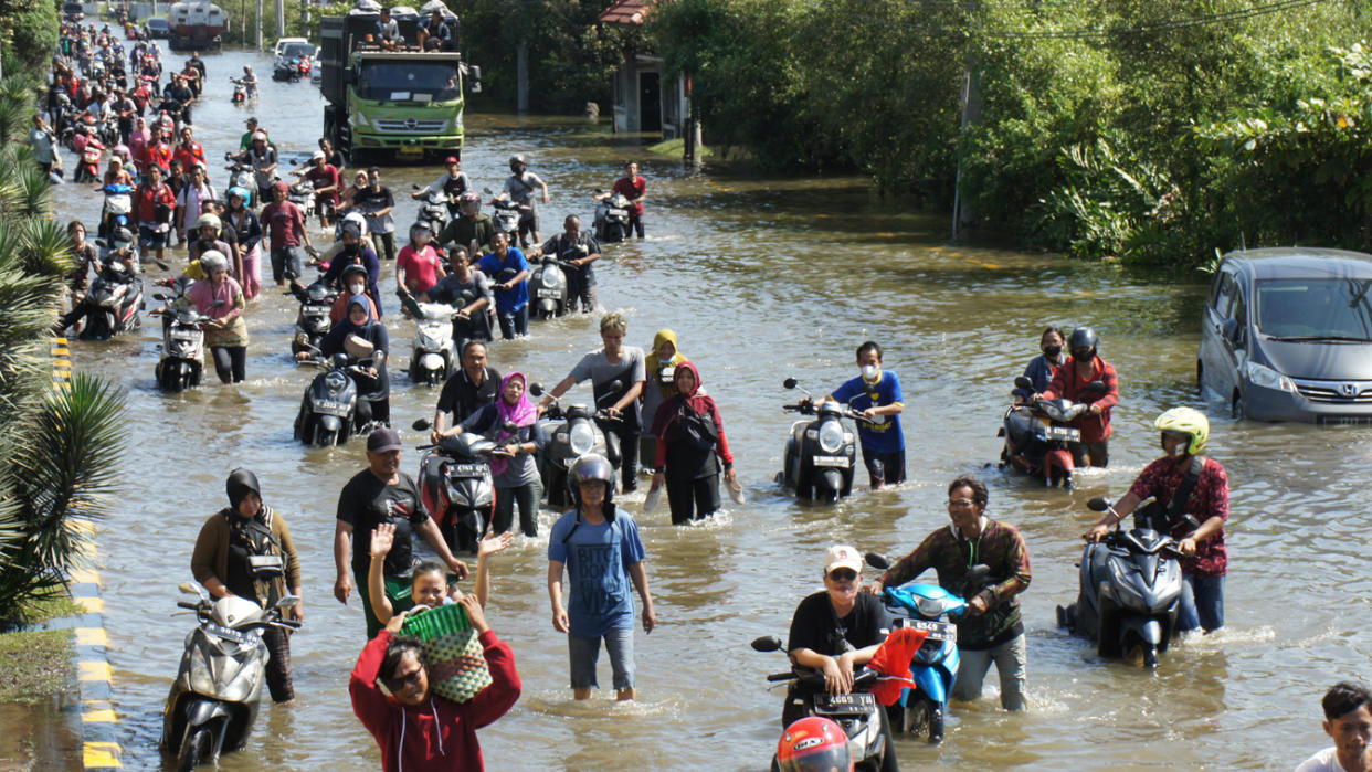 A crowd of people, many in helmets and pushing their motorcycles, wade down a road through floodwaters.