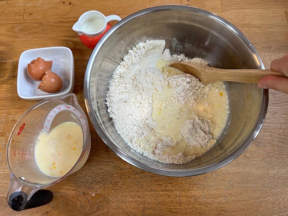 An overhead shot shows a hand stirring a mixture of egg and milk added to a bowl of flour, sugar, baking powder, and butter, with a wooden spoon.