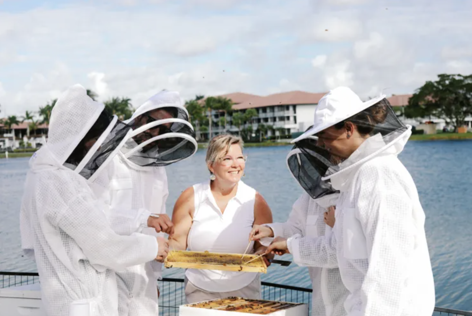 a woman holding a honeycomb and people around her in white protective bee outfits