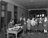 <p>A group of schoolchildren line up in an orderly fashion at their teacher's desk.</p>