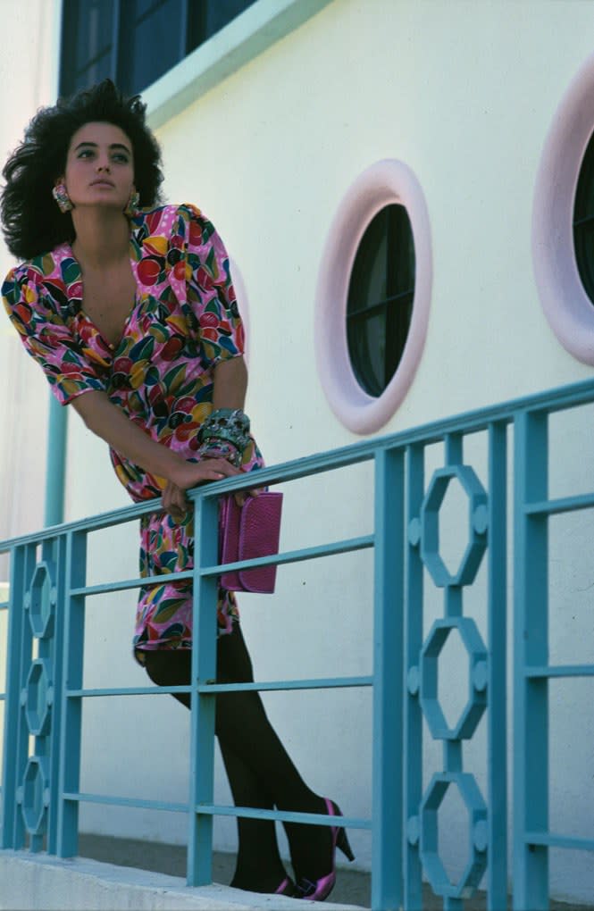 Model Josefina Toledo (née Josefina Victoria Toledo) leaning over a railing, holding a pink purse by Carlos Falchi and wearing a multicolored dress by Emanuel Ungaro, bracelets and earrings by Laurette Gneagy and black stockings by Evan-Picone Hosiery