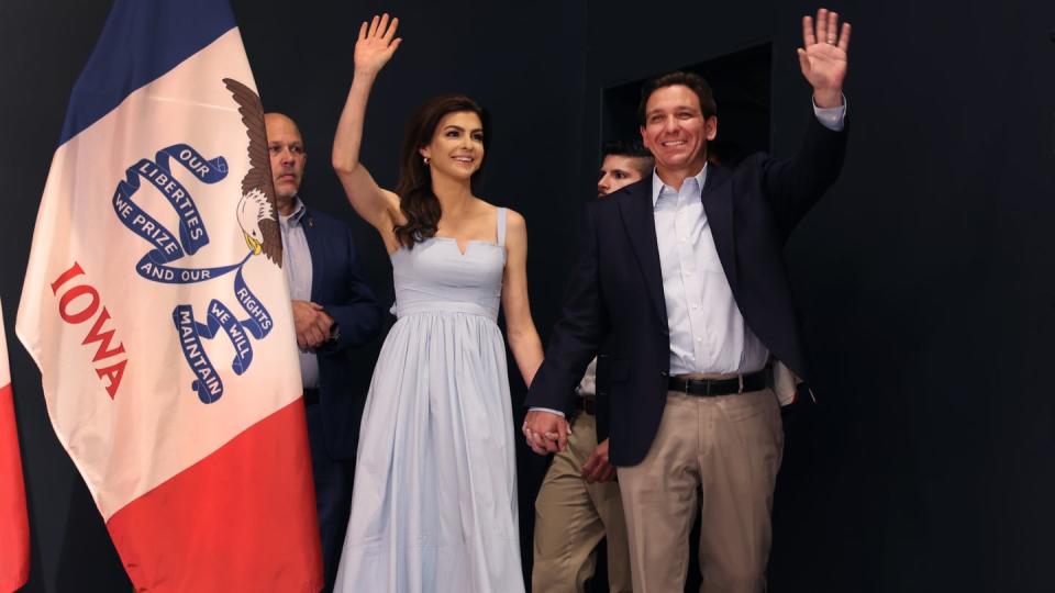 casey desantis and ron desantis hold hands, smile, and wave to a crowd out of view, she wears a baby blue dress and he wears a suit jacket with a light blue collared shirt and khaki pants