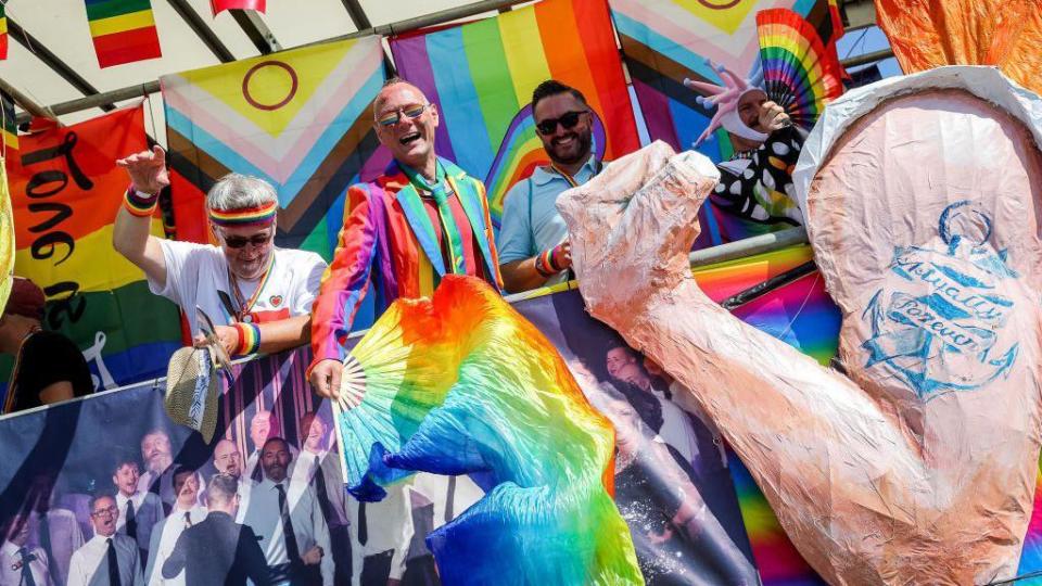 Three men in rainbow attire stand in front of pride flags