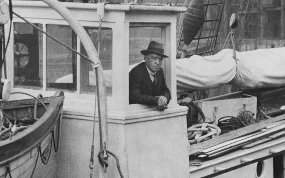 Shackleton wears a Trilby hat and leans out of the cabin of a ship in this black and white photo