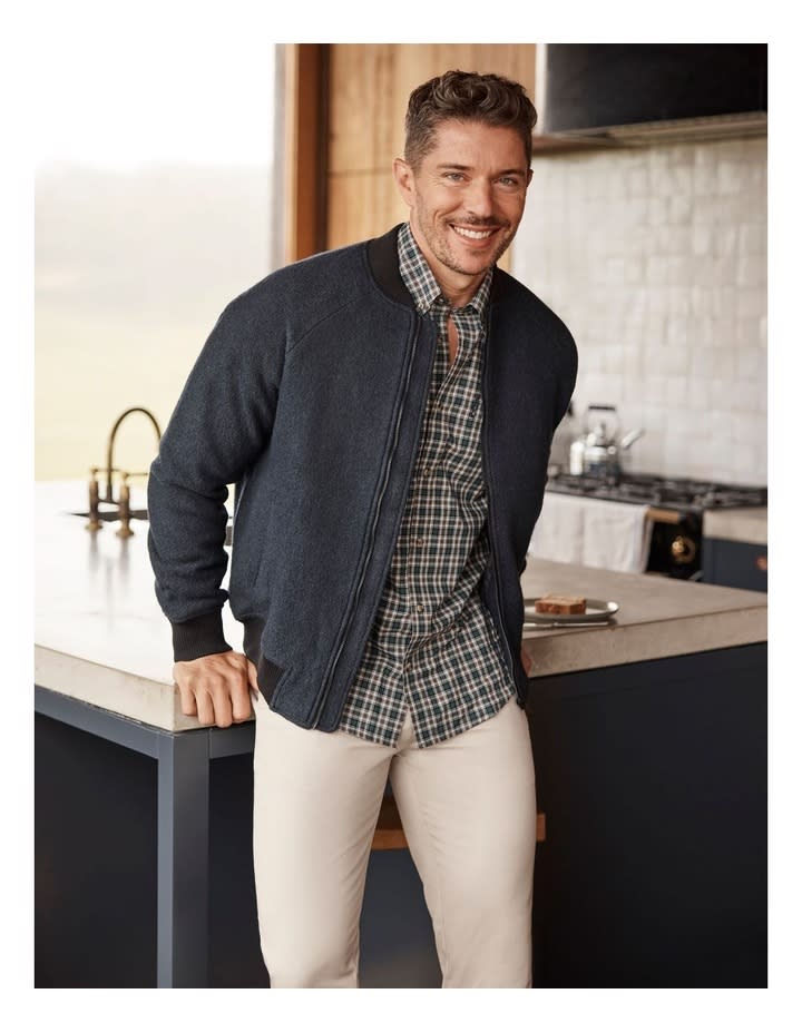 A man stands smiling resting his hands on a kitchen bench wearing white pants, tartan checked shirt and charcoal blazer