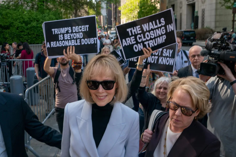 E. Jean Carroll, wearing dark glasses and a light blue suit, with her lawyer, pushes past anti-Trump protesters outside the court, carrying placards saying: Trump&#39;s Deceit Is Beyond a Reasonable Doubt and The Walls Are Closing In on Trump.