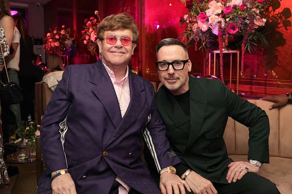 Elton John in a purple suit and David Furnish in a black suit sitting together at an indoor event with floral decorations in the background