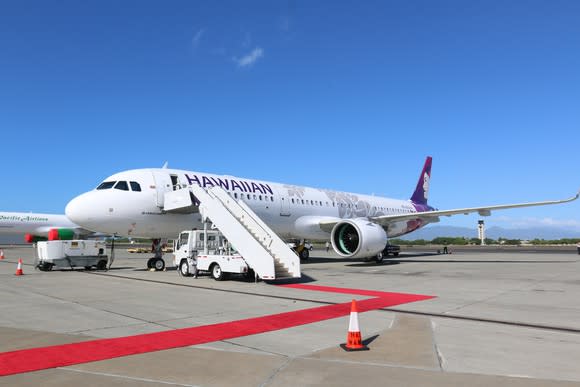 A red carpet sitting at the bottom of the stairs leading to a Hawaiian Airlines A321neo sitting on a tarmac under blue skies