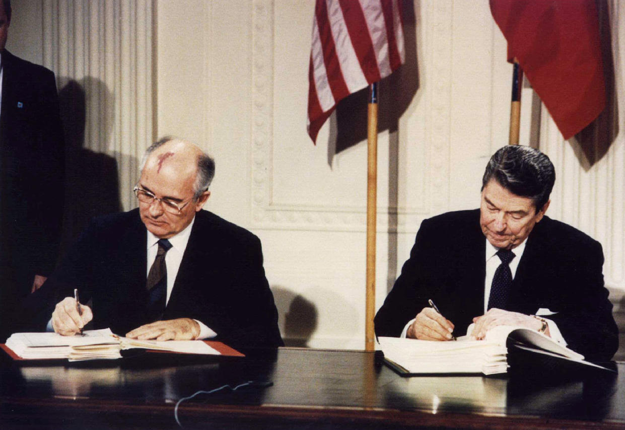 Mikhail Gorbachev and President Ronald Reagan, their nation's flags behind them, side by side at a desk, each sign a page in a huge file of papers.