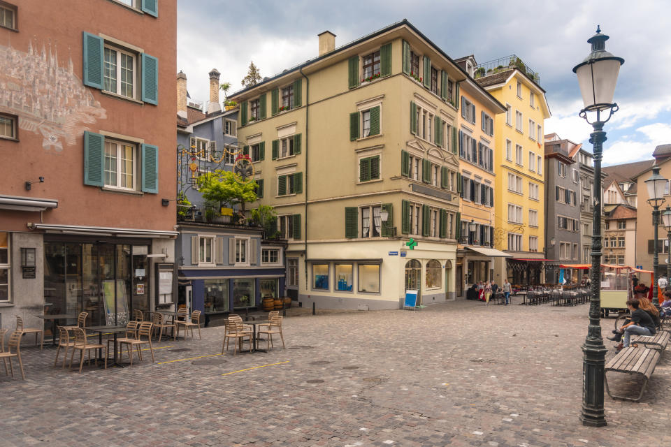 A cobblestone square in a European town is surrounded by pastel-colored buildings with outdoor seating. People are casually walking or sitting in the area