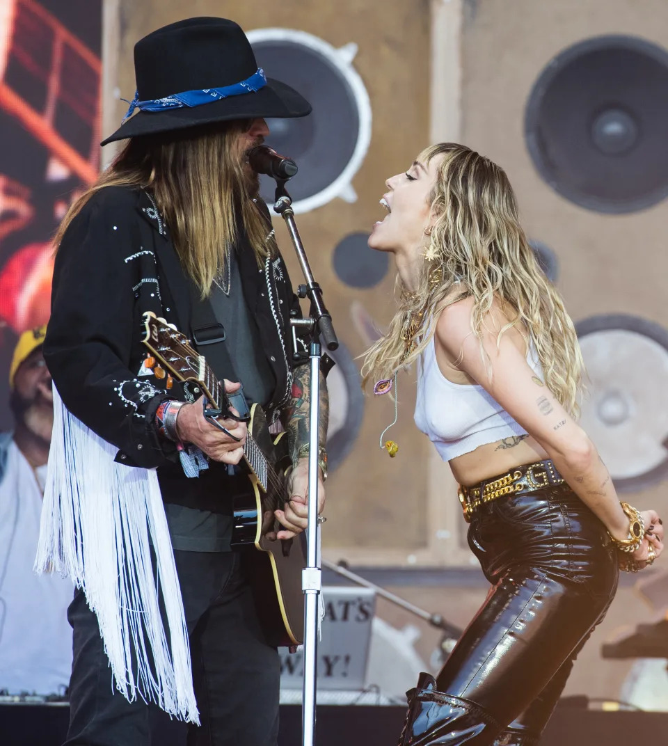 Billy Ray Cyrus and Miley Cyrus performing on stage. Billy Ray wears a fringed jacket and hat, while Miley is in a white crop top and black leather pants