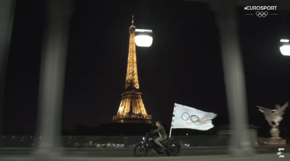 Person rides a motorcycle carrying an Olympic flag at night in front of the illuminated Eiffel Tower. Eurosport and Olympic logos are displayed on the upper right