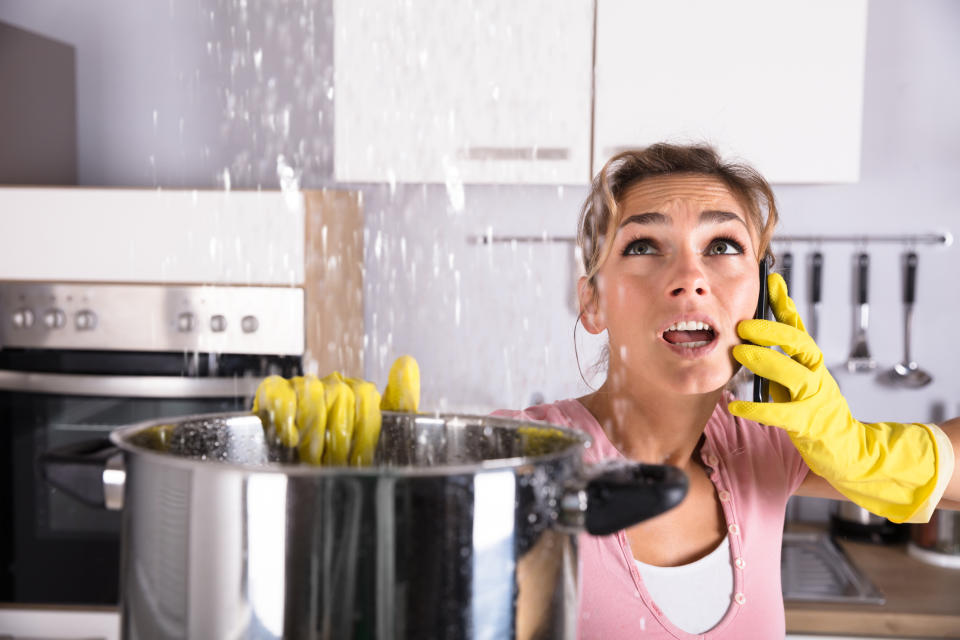 Woman who's wearing yellow plastic gloves on phone looking up as water pours down from the ceiling into a giant pot.