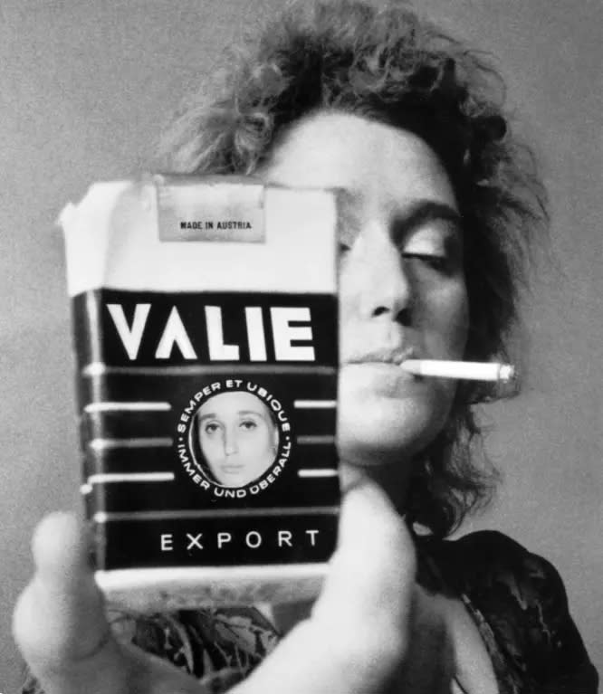 Smart Export, 1970, by Valie Export. A black-and-white photograph of a person smoking a cigarette, holding up a cigarette packet that obscures the right side of their face, reading 