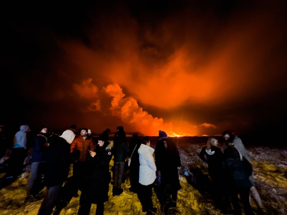 People watch as a volcano erupts in the distance with dark, reed skies.
