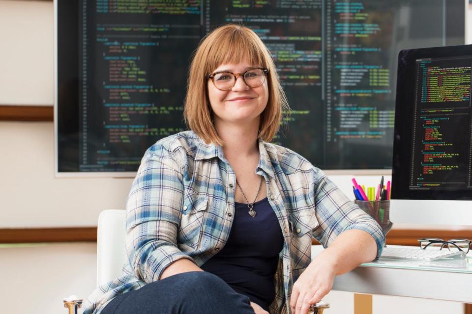 A woman sits in front of a monitor displaying computer code