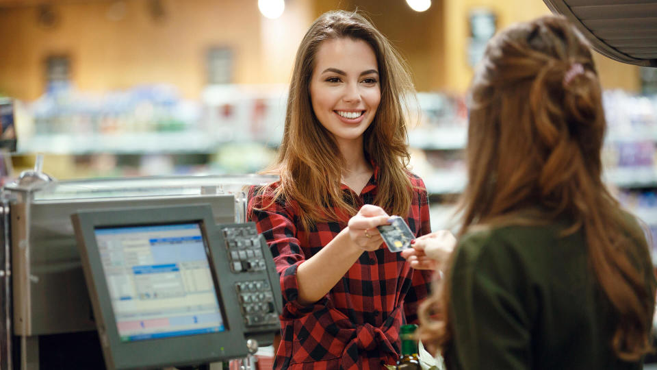 Image of smiling young lady standing in supermarket shop near cashier's desk holding credit card.