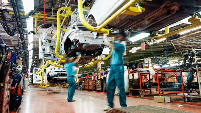 Two workers wearing green shirts and blue pants work underneath a car in an automobile factory.