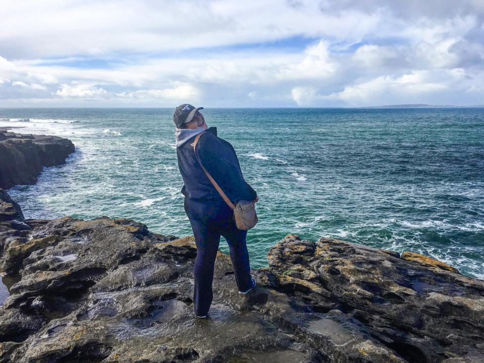 A woman in a baseball hat stands on rocks beside the ocean, looking back at the camera.