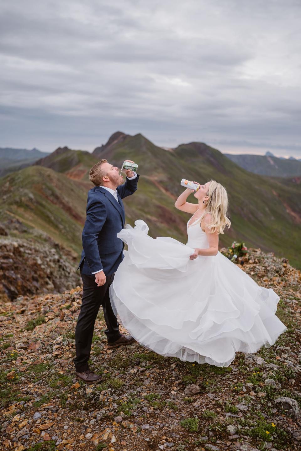 A groom and bride drink beers on a mountaintop.