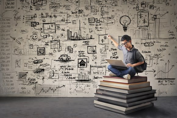 A man sitting on a stack of books points to a wall covered in scribbles of ideas.