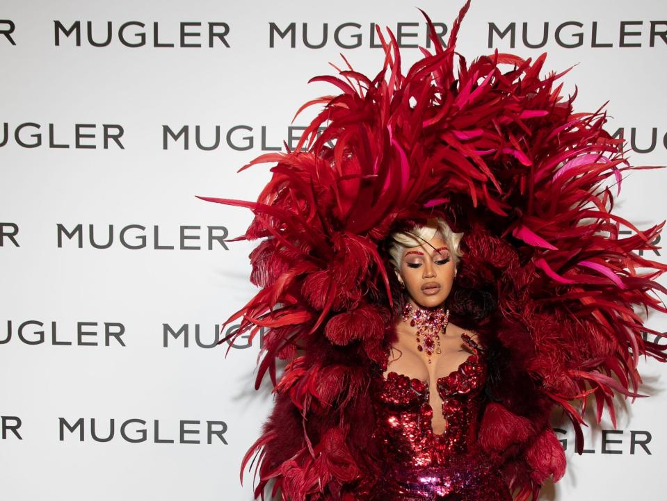 Rapper Cardi B in a red dress with red feathers and a red cape in front of a wall that reads "MUGLER"