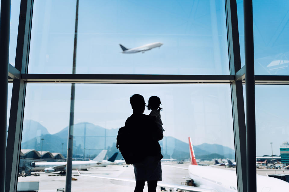 Silhouette eines Vaters mit Kind auf dem Arm, die gemeinsam ein startendes Flugzeug beobachten.