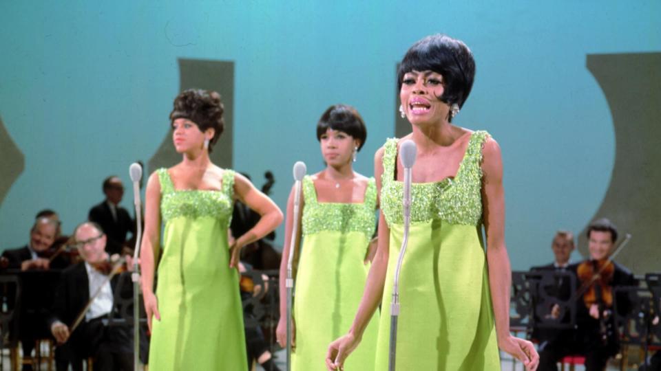 the supremes perform on stage in matching green scoopneck sleeveless gowns, behind them sit musicians