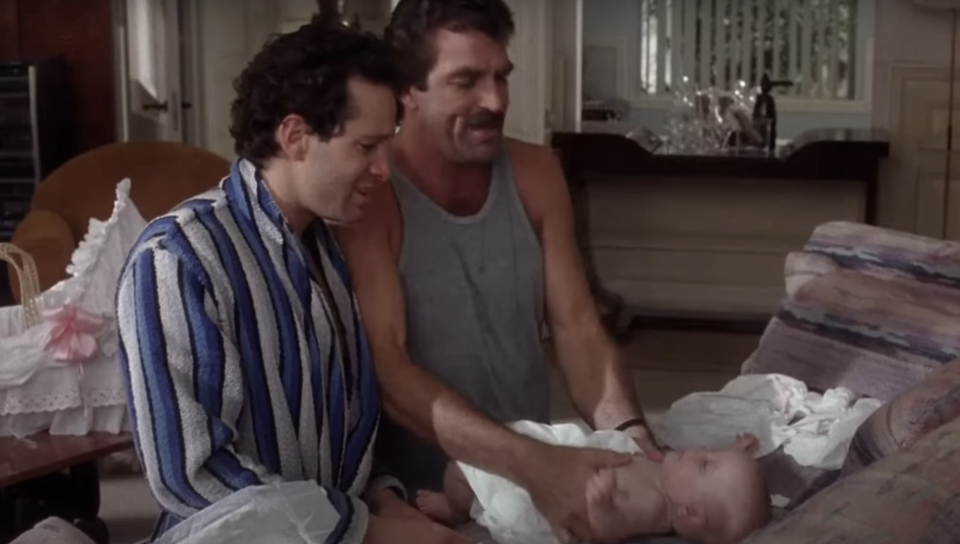 Two men, one in a striped robe and the other in a sleeveless shirt, gently attending to a baby on a couch