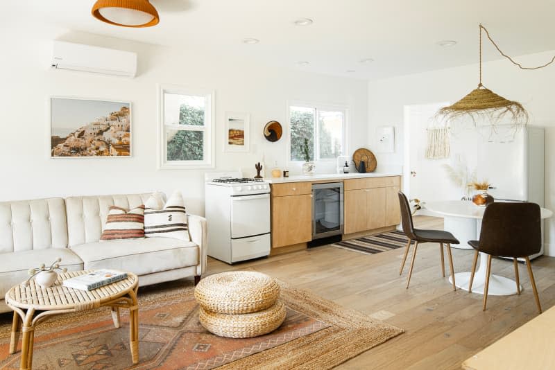 living, dining, and kitchen area with wood floors, wood kitchen cabinets, white stove, white dining table with black chairs, straw pendant light, pale neutral sofa with cane table, sisal or jute rug, straw pillows