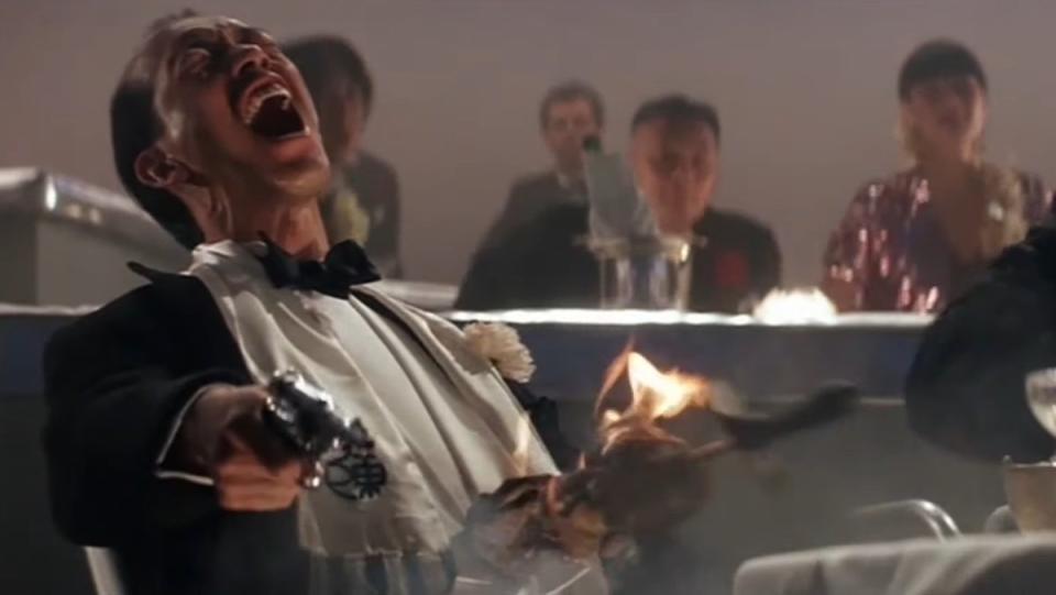 A man in a white tux fires a gun and screams with a flaming skewer of meat in his belly