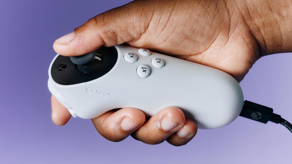 Die rechte Hand einer Person hält einen Xbox Accessibility Joystick vor einem violetten Hintergrund.