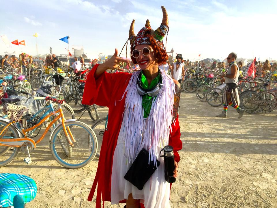 A Burning Man attendee wearing a red cloak, sunglasses, and a horned helmet in front of dozens of bikes on the playa.
